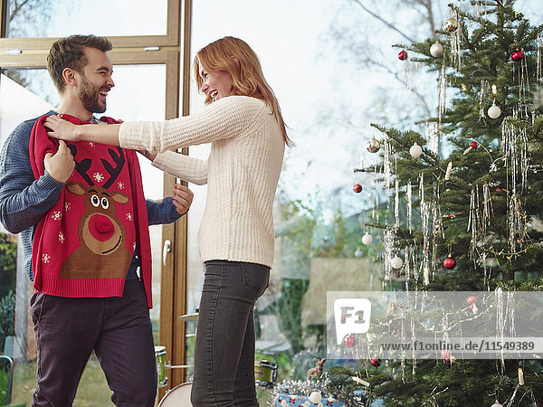 Paar steht vor dem Weihnachtsbaum und probiert den Weihnachtspullover an.