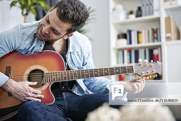 Young man sitting on couch tuning guitar