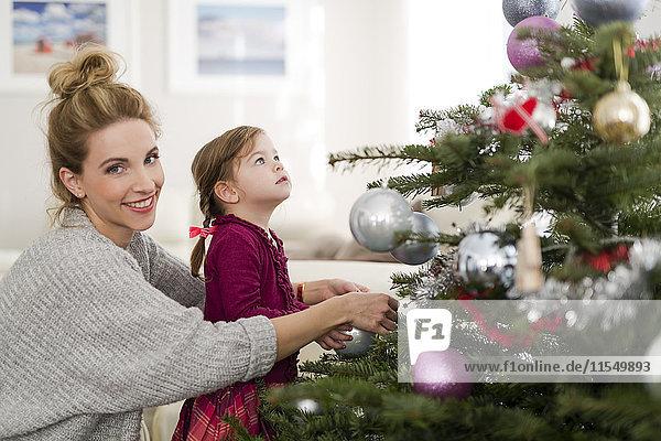 Porträt einer lächelnden Frau  die mit ihrer kleinen Tochter im Wohnzimmer den Weihnachtsbaum schmückt.