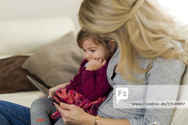 Kleines Mädchen sitzt auf dem Schoß ihrer Mutter und schaut auf ein digitales Tablett.