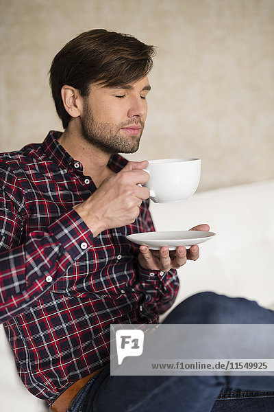 Porträt eines Mannes mit geschlossenen Augen  der auf einer Couch sitzt und eine Tasse Kaffee hält.