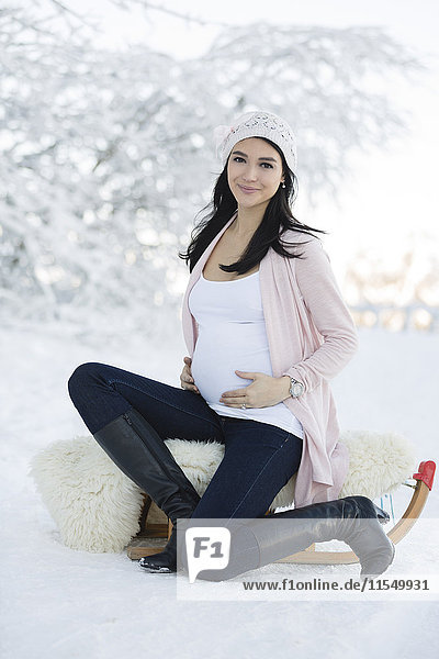 Lächelnde Schwangere im Freien mit Schlitten im Winter