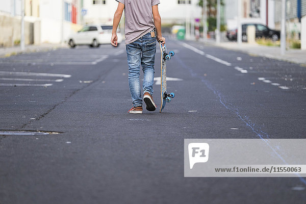 Rückansicht des Jungen  der sein Skateboard auf einer Straße trägt.