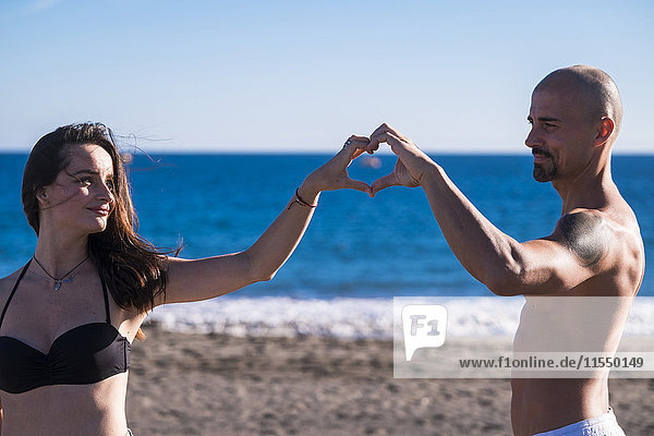 Spanien  Teneriffa  Porträt eines glücklichen Paares am Strand  das mit den Händen ein Herz baut.