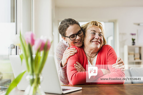 Smiling young woman hugging senior woman at table with laptop