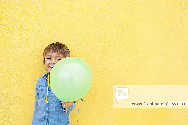 Lächelnder kleiner Junge mit grünem Ballon und Luftschlange vor gelber Wand