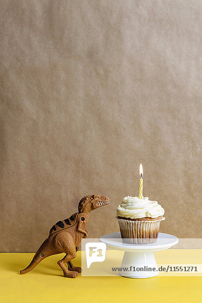 Spielzeugdinosaurier und Tassenkuchen mit angezündeter Kerze auf einem Kuchenständer
