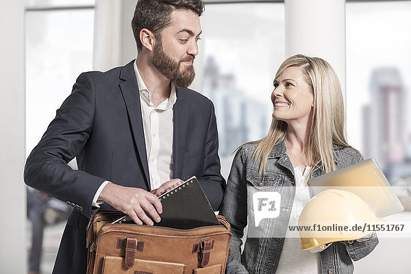 Mann mit Aktentasche und Frau mit Schutzhelm im Büro lächeln sich an.