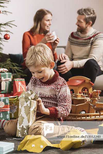Kleiner Junge freut sich über Weihnachtsgeschenk mit Eltern im Hintergrund