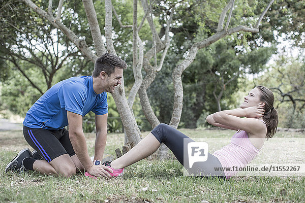 Young man assisting woman doing sit-ups on a meadow