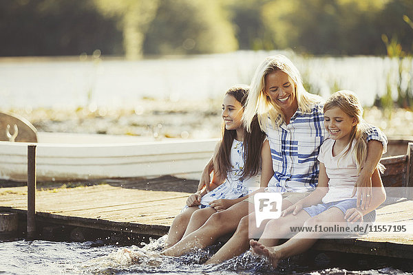 Smiling  affectionate mother and daughters on dock splashing feet in lake