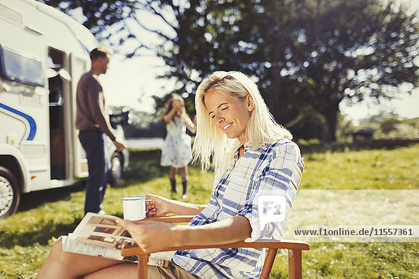 Frau liest Magazin und trinkt Kaffee draußen im sonnigen Wohnmobil