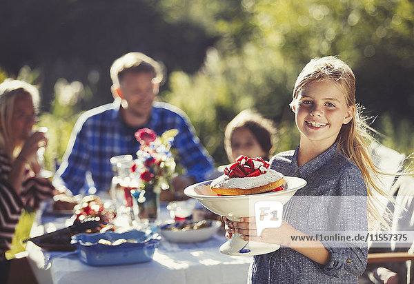 Portrait lächelndes Mädchen serviert Erdbeerkuchen für die Familie an einem sonnigen Gartenparty-Tisch.