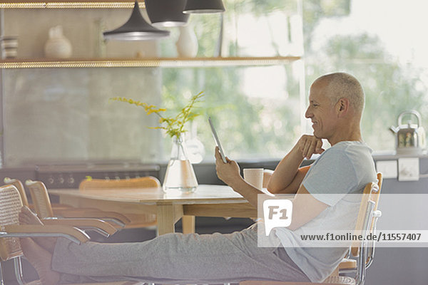 Älterer Mann  der sich entspannt und mit hochgelegten Füßen am Esstisch ein digitales Tablet benutzt