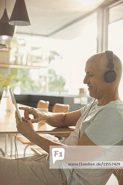 Älterer Mann  der mit Kopfhörern und digitalem Tablet am Esstisch Musik hört