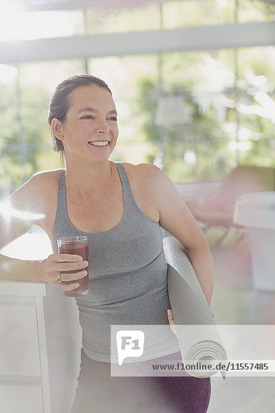 Lächelnde reife Frau mit Yogamatte trinkt gesunden Frucht-Smoothie