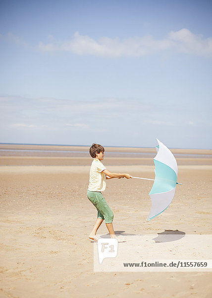 Wind zieht Regenschirm in den Händen eines Jungen am sonnigen Sommerstrand