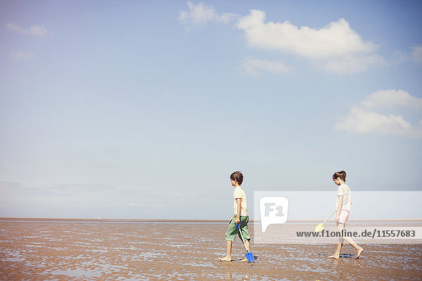 Bruder und Schwester mit Schaufeln im nassen Sand am sonnigen Sommerstrand unter blauem Himmel
