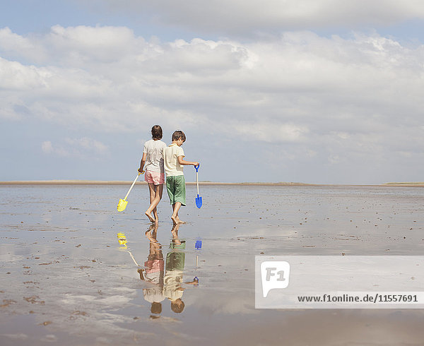 Bruder und Schwester gehen mit Schaufeln im nassen Sand am sonnigen Sommerstrand
