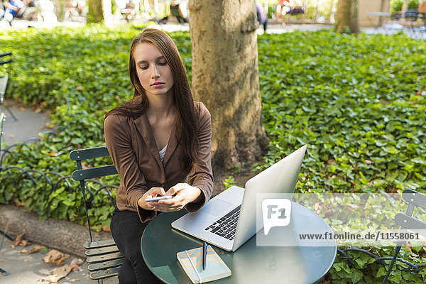 Junge Geschäftsfrau mit Laptop sitzt am Tisch in einem Park SMS