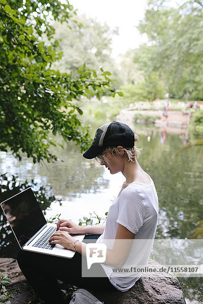 Junge Frau sitzt am Seeufer im Park mit Laptop