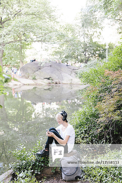 Junge Frau am Seeufer im Park mit Kopfhörer und Blick aufs Handy