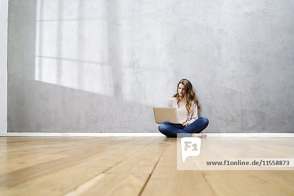 Blonde Frau auf dem Boden vor grauer Wand sitzend mit Laptop