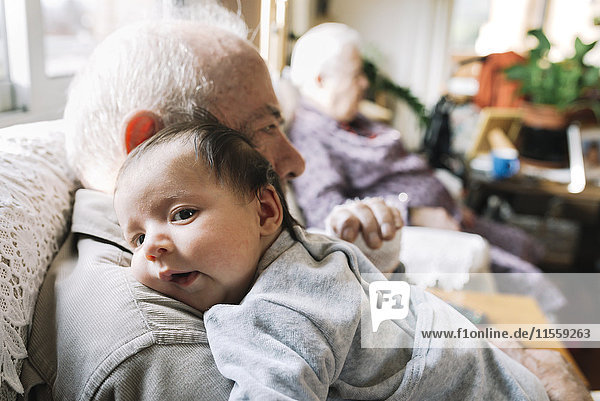 Great-grandfather holding baby at home with his wife in background
