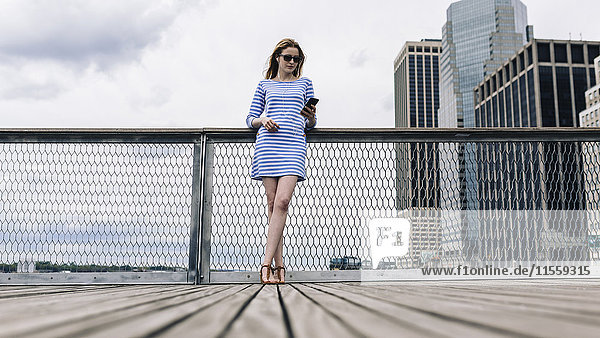 USA  New York City  Young woman standing in Manhattan at railing  holding smart phone