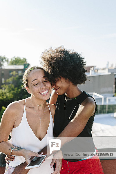 Female friends standing on rooftop  embracing and holding smart phones