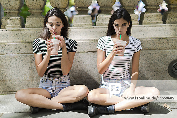 Zwei Zwillingsschwestern sitzen im Freien und trinken etwas zum Mitnehmen.