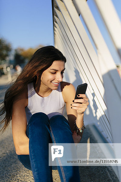 Lächelnde junge Frau schaut bei Sonnenuntergang aufs Handy