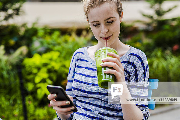 Young woman drinking a smoothie and checking cell phone outdoors