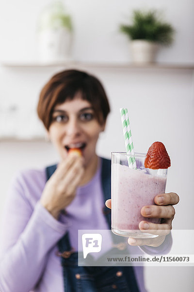 Frau hält ein Glas Erdbeer-Smoothie  während sie eine Frucht isst.