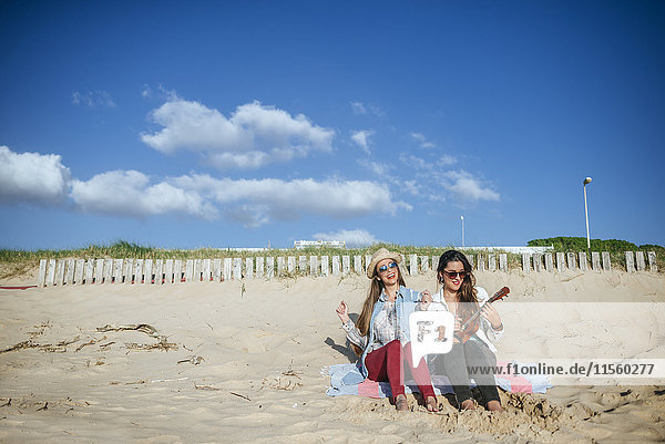 Zwei junge Frauen mit Ukulelele sitzen auf einer Decke am Strand.