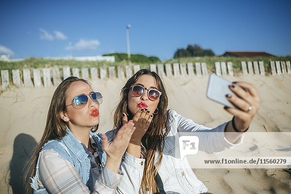 Two young women taking selfie on the beach while blowing a kiss