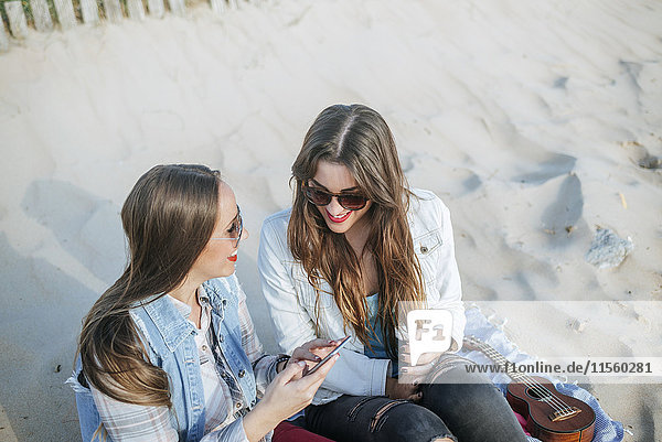 Two young women sitting on the beach looking using cell phone