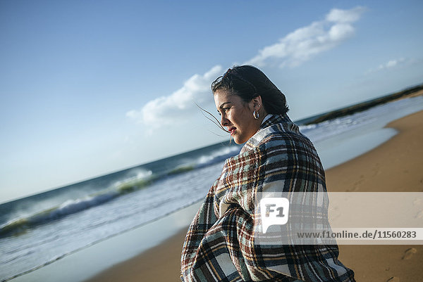 Junge Frau mit Decke am Strand mit Blick auf das Meer
