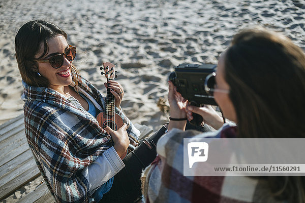 Smiling young woman playing ukulele while her friend recording with video camera