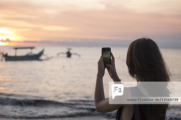 Indonesien  Bali  Frau beim Fotografieren des Sonnenuntergangs über dem Meer
