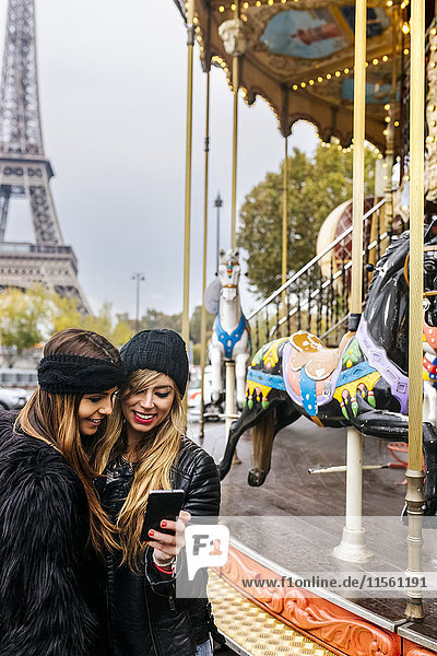 France  Paris  two best friends using their smartphone at a carousel with the Eiffel Tower in the background