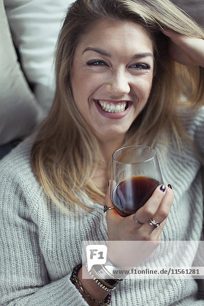 Porträt einer lachenden blonden Frau mit einem Glas Kaffee auf der Couch zu Hause.