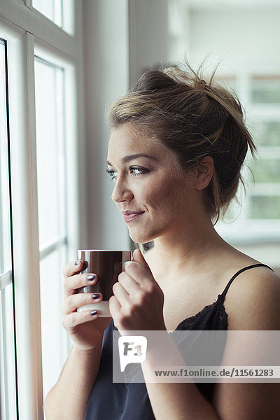 Porträt einer lächelnden blonden Frau mit einem Glas Kaffee durchs Fenster schauend