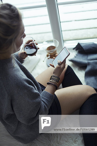 Junge Frau mit Tasse Kaffee entspannt auf Schaffell zu Hause Blick auf Handy