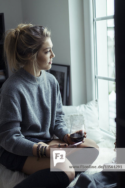 Young woman with cup of coffee and smartphone looking through window
