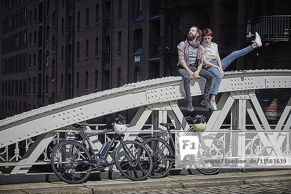 Deutschland  Hamburg  Paar mit Elektrofahrrädern auf Brückengeländer sitzend