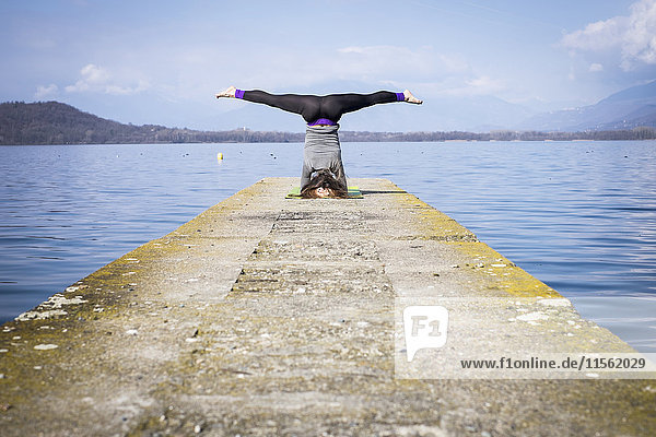 Frau  die Yoga praktiziert und einen Kopfstand auf einem Pier am See macht.