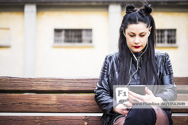Dunkelhaarige junge Frau sitzt auf der Bank und hört Musik mit Kopfhörern und Smartphone.