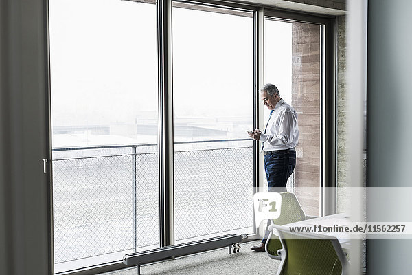 Senior Manager im Büro stehend am Fenster mit Smartphone