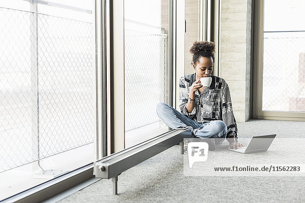 Young woman sitting on floor using laptop  drinking coffee